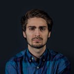 A thoughtful young man in a blue checked shirt looking serious against a dark background.
