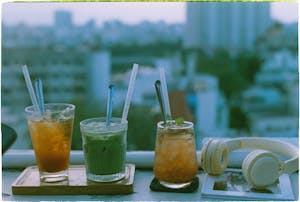 Three vibrant summer drinks with straws on a tray beside headphones, against an urban skyline backdrop.